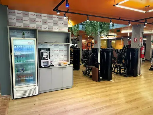 Geladeira de bebidas e máquina de café instaladas dentro do salão, ao lado das estações de musculação.