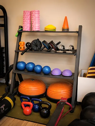 Estante com rolos de liberação rosa, cones, rodinhas de abdominal, bolas azuis, meias-bolas lilás, dois bosus laranja e kettlebells no chão.